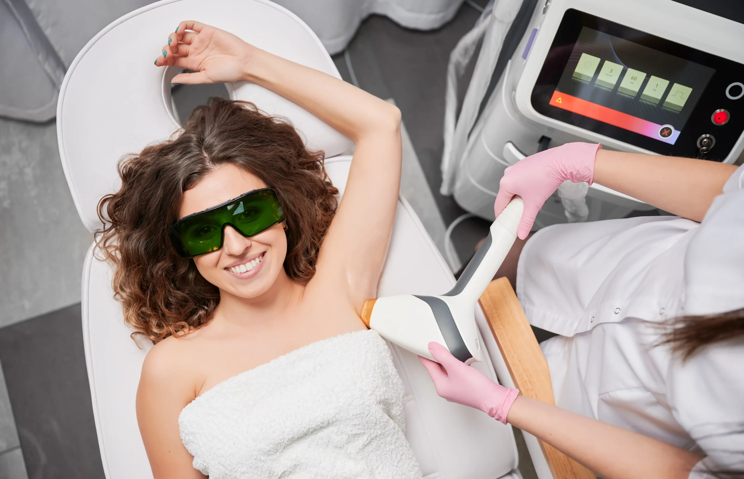 A smiling woman wearing protective goggles receiving underarm laser hair removal treatment